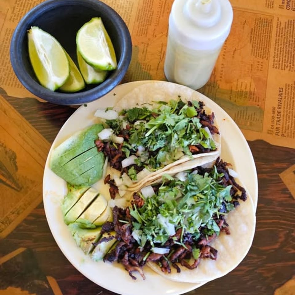 Two Los Vega Mexican Restaurant tacos with meat, cilantro, onions, and avocado slices, with limes and sauce.
